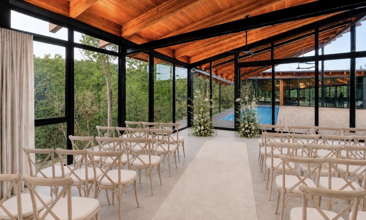 Pool House ceremony — cross-back chairs and florals framed by glass walls and pool views